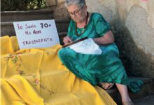Una donna over 70 ricama seduta sui gradini accanto al cartello "Io sono 70+ ma non trasparente"