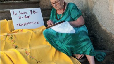 Una donna over 70 ricama seduta sui gradini accanto al cartello "Io sono 70+ ma non trasparente"