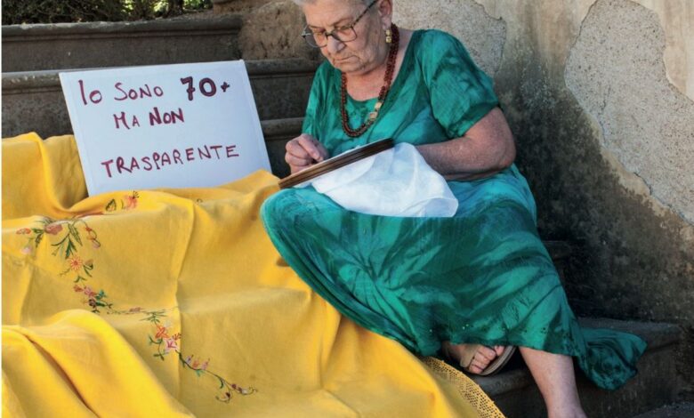 Una donna over 70 ricama seduta sui gradini accanto al cartello "Io sono 70+ ma non trasparente"